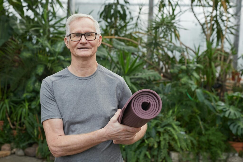 Smiling senior man with gray hair holding a yoga mat in a lush greenhouse.